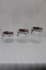 Three transparent acrylic airtight storage jars of varying sizes featuring wooden lids, metal clamp locks, and attached spoons, displayed neatly on a white kitchen countertop from One More Crockery.