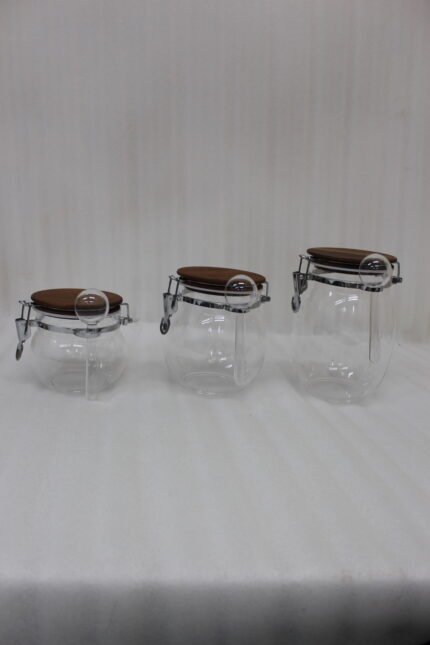 Three transparent acrylic airtight storage jars of varying sizes featuring wooden lids, metal clamp locks, and attached spoons, displayed neatly on a white kitchen countertop from One More Crockery.