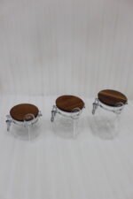 Three transparent acrylic airtight storage jars of varying sizes featuring wooden lids, metal clamp locks, and attached spoons, displayed neatly on a white kitchen countertop from One More Crockery.