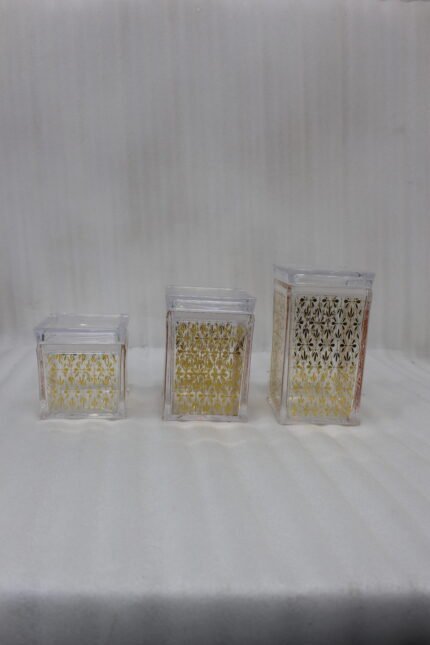 Set of three clear acrylic kitchen storage jars with elegant gold patterned designs, displayed on a marble countertop.