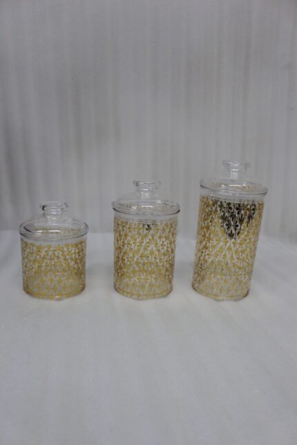 Three clear acrylic storage jars of varying heights featuring a decorative gold pattern and matching lids, displayed on a kitchen countertop next to spices.