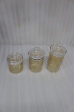 Three clear acrylic storage jars of varying heights featuring a decorative gold pattern and matching lids, displayed on a kitchen countertop next to spices.