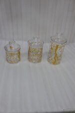 Three transparent acrylic storage jars of varying heights featuring a decorative gold branch pattern, arranged on a marble kitchen countertop with plates and a plant in the background.