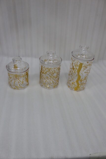 Three transparent acrylic storage jars of varying heights featuring a decorative gold branch pattern, arranged on a marble kitchen countertop with plates and a plant in the background.