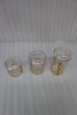 Three transparent acrylic storage jars of varying heights featuring a decorative gold branch pattern, arranged on a marble kitchen countertop with plates and a plant in the background.