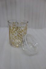 Three transparent acrylic storage jars of varying heights featuring a decorative gold branch pattern, arranged on a marble kitchen countertop with plates and a plant in the background.