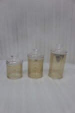Three luxury clear acrylic jars with gold horizontal stripe patterns and matching lids displayed on a marble countertop in a modern kitchen.