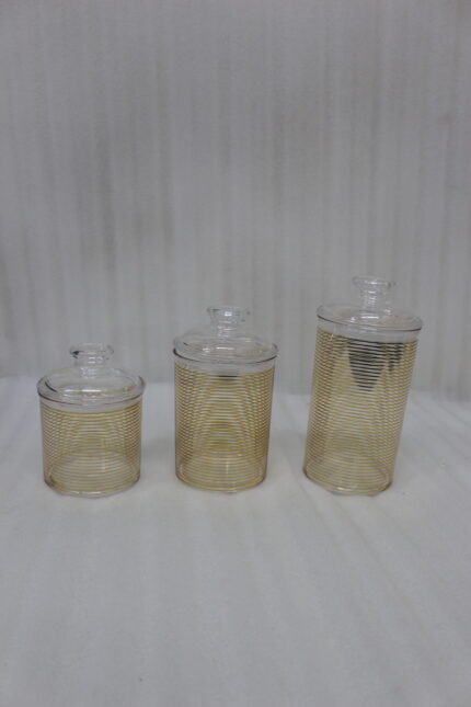 Three luxury clear acrylic jars with gold horizontal stripe patterns and matching lids displayed on a marble countertop in a modern kitchen.