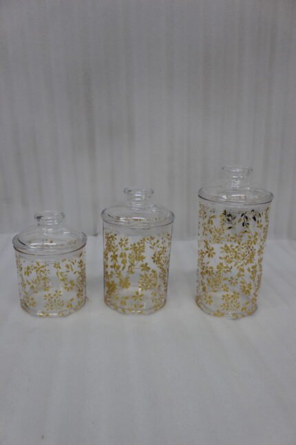 Three clear acrylic jars of varying heights featuring a gold floral print and clear lids, arranged on a marble kitchen countertop.
