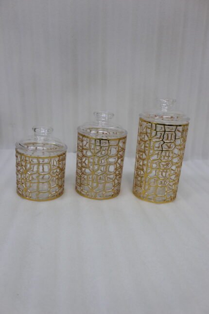 Three luxurious clear acrylic jars of varying heights featuring intricate gold geometric designs and matching lids, displayed neatly on a white marble countertop with a small golden spoon.