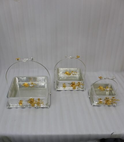 Three silver textured decorative serving dishes with gold floral accents and overhead arched handles on a wooden coffee table.