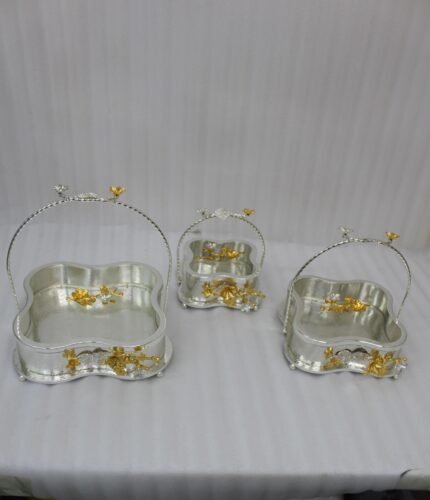 Three-piece silver hammered decorative bowl set with gold floral accents and overhead handles arranged on a wooden table.