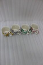 Four ceramic coffee mugs with colorful floral patterns in yellow, purple, green, and pink lined up on a marble countertop.