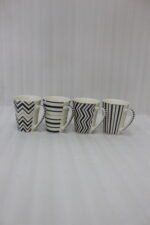 Set of four modern ceramic coffee mugs featuring black and white geometric, chevron, and striped patterns with gold rim accents on a marble countertop.