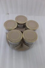 Four modern 6-piece revolving glass spice jar sets with gold trim lids displayed on a white kitchen countertop in beige, cream, black, and white colors.