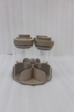 Four-piece clear glass spice jar sets with airtight lids displayed on rotating organizer trays in black, beige, tan, and white on a marble kitchen countertop.