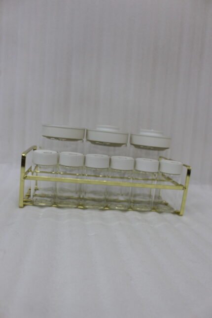 9-piece clear glass spice jar set with white and gold airtight lids arranged on a gold metal rack on a kitchen countertop.