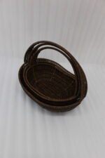Set of three handwoven dark brown oval rattan baskets with handles resting on a light wooden table.