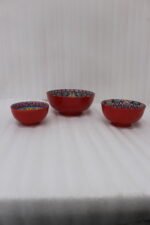 A set of three decorative ceramic bowls in a kitchen. They have rich red exterior glazes and detailed, traditional hand-painted Moroccan-style patterns in blue, yellow, and red on the inside, sitting on a wooden counter next to spice jars.