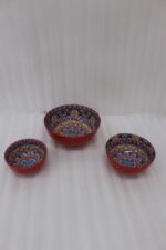 A set of three decorative ceramic bowls in a kitchen. They have rich red exterior glazes and detailed, traditional hand-painted Moroccan-style patterns in blue, yellow, and red on the inside, sitting on a wooden counter next to spice jars.