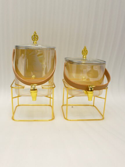 Two ribbed glass beverage dispensers with gold spigots, gold metal stands, decorative lids, and brown leather carrying straps on a marble kitchen counter.