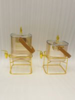 Two ribbed glass beverage dispensers with gold spigots, gold metal stands, decorative lids, and brown leather carrying straps on a marble kitchen counter.