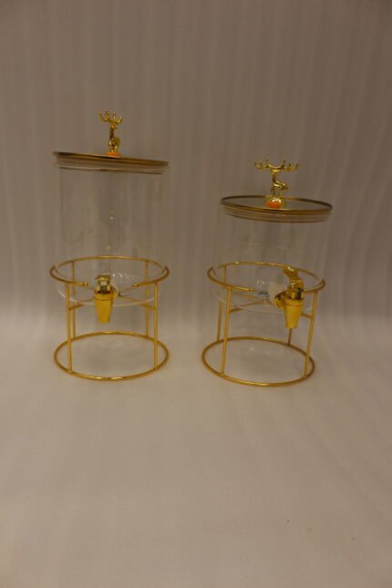 Two luxury clear glass beverage dispensers with gold deer lid handles and matching metallic stands on a marble countertop