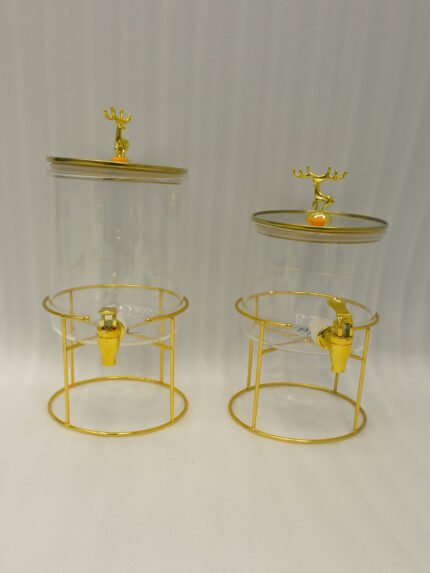Two luxury clear glass beverage dispensers with gold deer lid handles and matching metallic stands on a marble countertop