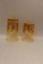 Two elegant clear glass drink dispensers with fruit prints, gold spigots, and gold deer lid handles resting on matching metal wire stands.