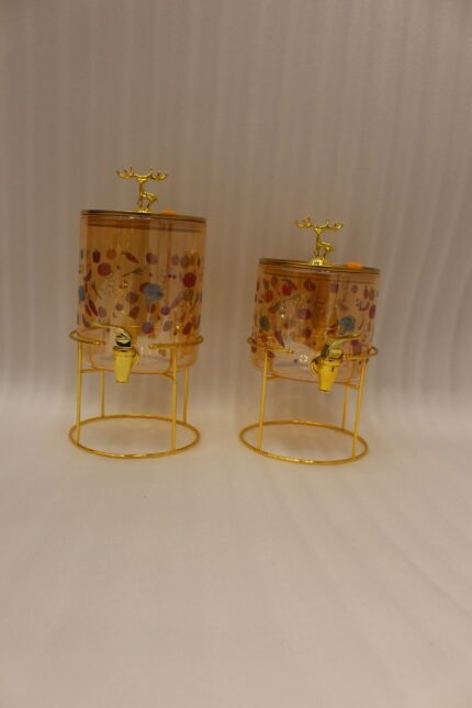 Two elegant clear glass drink dispensers with fruit prints, gold spigots, and gold deer lid handles resting on matching metal wire stands.