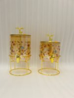 Two elegant clear glass drink dispensers with fruit prints, gold spigots, and gold deer lid handles resting on matching metal wire stands.