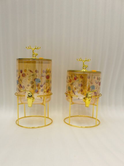 Two elegant clear glass drink dispensers with fruit prints, gold spigots, and gold deer lid handles resting on matching metal wire stands.