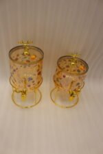 Two elegant clear glass drink dispensers with fruit prints, gold spigots, and gold deer lid handles resting on matching metal wire stands.