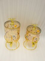 Two elegant clear glass drink dispensers with fruit prints, gold spigots, and gold deer lid handles resting on matching metal wire stands.