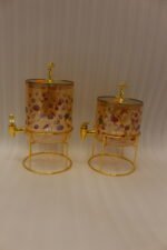 Two elegant clear glass drink dispensers with fruit prints, gold spigots, and gold deer lid handles resting on matching metal wire stands.