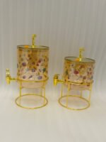 Two elegant clear glass drink dispensers with fruit prints, gold spigots, and gold deer lid handles resting on matching metal wire stands.