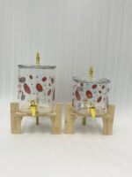 Two strawberry pattern glass drink dispensers with gold spigots set on wooden stands in a bright kitchen context.