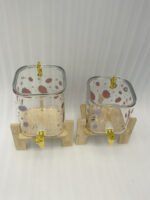 Two strawberry pattern glass drink dispensers with gold spigots set on wooden stands in a bright kitchen context.