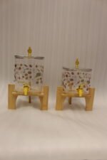 Two square glass beverage dispensers with fruit prints, gold spigots, and wooden stands on a kitchen counter