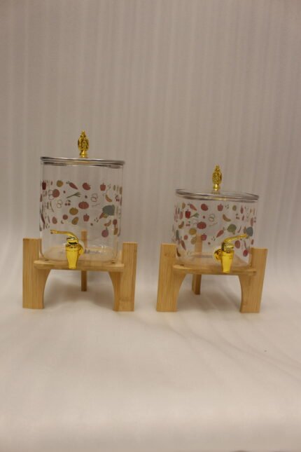 Two square glass beverage dispensers with fruit prints, gold spigots, and wooden stands on a kitchen counter
