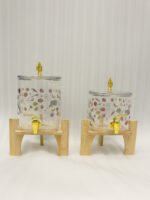 Two square glass beverage dispensers with fruit prints, gold spigots, and wooden stands on a kitchen counter