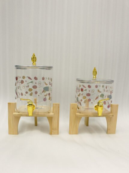 Two square glass beverage dispensers with fruit prints, gold spigots, and wooden stands on a kitchen counter