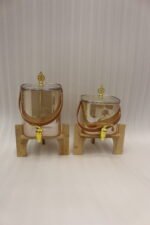 Two amber-shaded glass beverage dispensers with gold spigots, leather handles, and wooden stands on a kitchen counter.