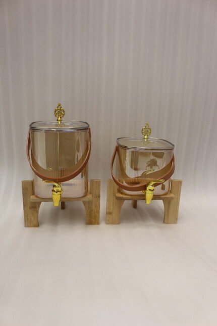Two amber-shaded glass beverage dispensers with gold spigots, leather handles, and wooden stands on a kitchen counter.