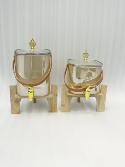 Two amber-shaded glass beverage dispensers with gold spigots, leather handles, and wooden stands on a kitchen counter.