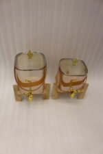 Two amber-shaded glass beverage dispensers with gold spigots, leather handles, and wooden stands on a kitchen counter.