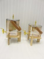 Two amber-shaded glass beverage dispensers with gold spigots, leather handles, and wooden stands on a kitchen counter.