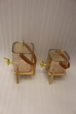 Two amber-shaded glass beverage dispensers with gold spigots, leather handles, and wooden stands on a kitchen counter.