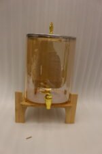 Amber glass drink dispenser with gold spigot resting on a natural wooden stand on a kitchen counter.