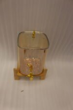 Amber glass drink dispenser with gold spigot resting on a natural wooden stand on a kitchen counter.